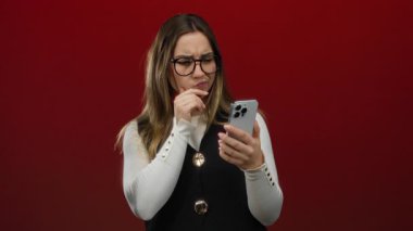 Gözlüklü genç kadın akıllı telefonuna canlı kırmızı bir arka planda bakarken düşünceli bir düşünce ve merak ifadesi sergiliyor..