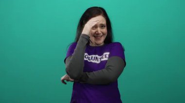 Kahverengi saçlı bir kadın gönüllü gömleğiyle yeşil arka plana gülüyor. Heyecan ve hizmete adanmışlığı simgeliyor.