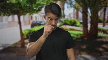 Genç İspanyol bir adam yeşil açık hava parkında jest yapıyor. Arka planda ağaçlar ve binalar var. Odaklanma ve dikkat hissi veriyor..
