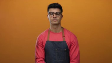 Gözlüklü ve önlüklü genç adam sağlam turuncu bir arka plana karşı duruyor, sıradan bir ortamda şık ve profesyonel bir aura yayıyor..