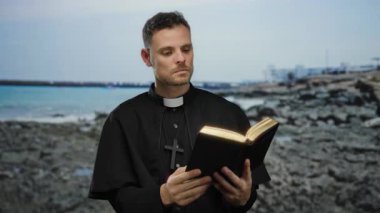 Papaz kayalık sahilinde açık bir kitapla kitap okuyor, sakin sahil atmosferini yakalıyor..