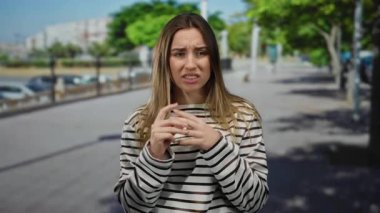 Çizgili tişört giyen kadın, arka planda yeşil ağaçlar ve binalar olan güneşli şehir bölgesinde sokakta endişeli görünüyor..