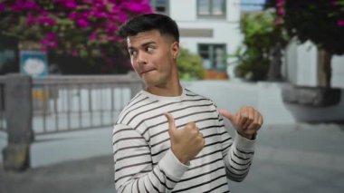 Gülümseyen ve el kol hareketi yapan genç bir adam canlı bir şehir caddesinde çiçekli bir arka planda gün ortasında çekilen çekimlerde neşe ve pozitiflik saçan.