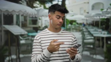 Terasta telefon tutan genç İspanyol bir adam şehir kafesinin dışında açık havada masalar ve sandalyelerle çevrili çizgili bir gömlek içinde dalgın dalgın görünüyor.