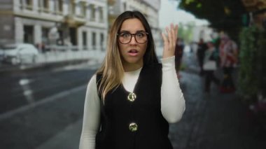 Genç bir kadın hareketli bir şehir ortamında şaşkınlığını dile getiriyor. Açık bir şehirde sarı saçlar, gözlükler ve günlük kıyafetler sergiliyor..