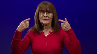 Kıdemli İspanyol kadın mavi bir arka plana gülümsüyor kırmızı bir süveter ve gözlük takıyor, dişlerini iki eliyle gösteriyor, pozitifliği ve sıcaklığı somutlaştırıyor.