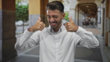 Beyaz tişörtlü bir adam taştan kolonlar ve kemerlerle kaplı bir caddede baş parmağını kaldırıyor..