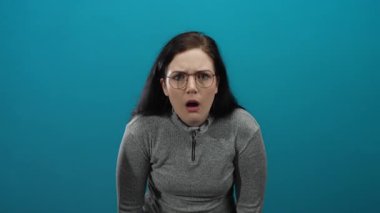 Gri kazaklı kadın, canlı mavi bir arka plan karşısında şaşkınlığını ifade ediyor. Şok, şaşkınlık, duygu ve yoğunluğu yakın çekimde gösteriyor..