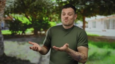 Parkta dövmeli bir adam yeşil gömlek giyerken meraklı görünüyor. Etrafı ağaçlarla çevrili. Güneş ışığı ve bulanık bir arka plan.