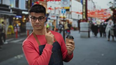 Gözlüklü genç adam hareketli şehir caddesinde kahve fincanı tutuyor. Etrafı canlı şehir dekorları ve çeşitli kalabalıklarla çevrili..
