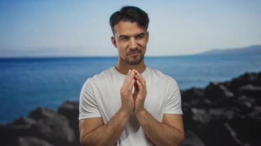 Deniz kenarındaki genç adam, derin düşüncelere dalmış, berrak mavi gökyüzünün altında sakin bir plaj arka planına karşı günlük beyaz gömlek giyiyor..