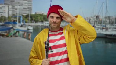 Sarı yağmurluklu ve kırmızı bereli genç adam deniz kıyısında oltayla selamlıyor, etrafı tekne ve suyla çevrili, neşeli ve deniz havasını iletiyor..