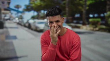 Kırmızı kazaklı genç adam güneşli bir şehir caddesinde gülümsüyor, rahat ve rahat bir atmosfer sergiliyor..