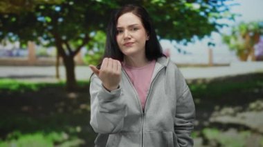 Parktaki bir kadın, yeşil ağaçlarla çevrili gri kapüşonlu bir kapüşonlu giysiyle kameraya doğru gülümsüyor..