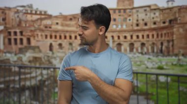 Mavi gömlekli genç İspanyol adam, açık şehir ortamındaki antik Roma harabelerinin perde arkasını düşünceli bir şekilde işaret ediyor..