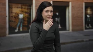 Açık havada gülümseyen esmer bir kadın sokakta dikiliyor siyah gömlek giyiyor, tuğla arka plan duygu ifadesi takınıyor.