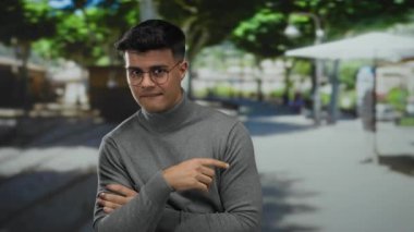 Gözlüklü genç adam güneşli bir şehir sokağında, açık şehir ortamında yeşil ağaçlarla çevrili neşeli bir ifadeyle kendini işaret ediyor..