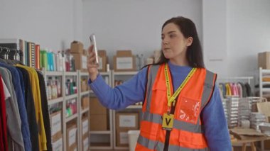 Gönüllü olarak selfie çeken bir kadın turuncu yelek giyiyor. Etrafı renkli kıyafetler ve kutularla çevrili..