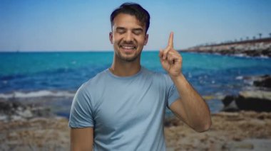 Deniz kenarındaki bir sahilde gülümseyen İspanyol genç adam, arka planda okyanus ve mavi gökyüzünü göstererek, keyif ve rahatlık hissini aktarıyor..