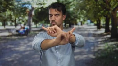 Yüzünde ciddi bir ifade olan genç adam yeşil bir açık hava parkında dur hareketi yapıyor..