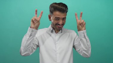 Beyaz gömlekli İspanyol genç adam iki eliyle turkuaz stüdyoda çifte barış işareti gösteriyor..