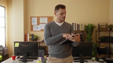 Ofisteki genç adam elinde tabletle bilgisayar ve bitkilerle duruyor. Profesyonel ve odaklanmış görünüyor..