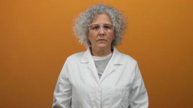 Beyaz önlüklü, gri saçlı ve gözlüklü bir kadın turuncu bir duvarın arkasında duruyor. Bilim ve sağlık alanında profesyonellik ve uzmanlık yayıyor..