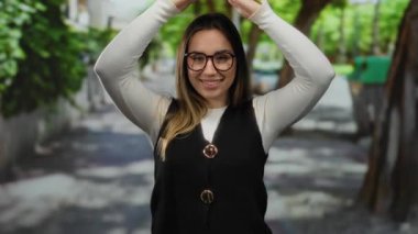 Yeşil ağaçlarla çevrili, gözlüklü ve siyah yelek giyen bir kadın, güneş ışığı altında poz verirken neşeli bir ifade sergiliyor..