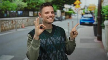 Kenar mahallelerde, yeşilliklerde ve arka planda gün ışığında görülebilen yol işaretleriyle şehir caddesinde gülümseyen bir adam iyi bir jest yapıyor..