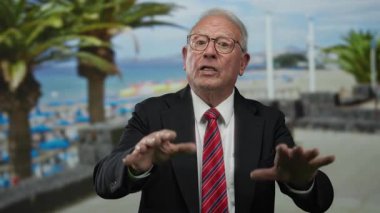 Takım elbiseli kıdemli bir adam deniz kenarında palmiye ağaçları ve okyanus manzarasıyla el kol geziyor. Dışarıya iş mesajını sakin ve profesyonel bir ortamda iletiyor..