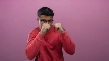 Kırmızı gömlekli genç adam canlı pembe bir arka plana karşı, gözlük takıyor ve oyuncu bir boks duruşu sergiliyor..