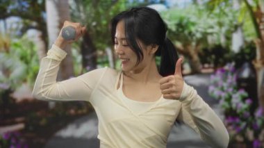 Genç Çinli bir kadın, yemyeşil bir parkta halterlerle egzersiz yapıyor. Parlak bir şekilde gülümsüyor ve başparmağını kaldırıyor. Hareketli ve enerjik bir anı yakalıyor..