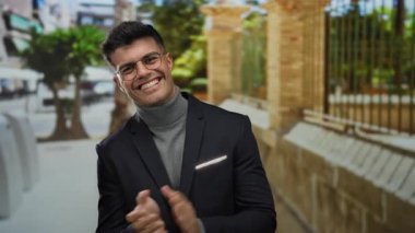 Gözlüklü genç İspanyol adam, bir şehir caddesinde gülümsüyor ve alkışlıyor, balıkçı yaka ve ceket giyiyor güneşli bir şehir zeminine karşı.