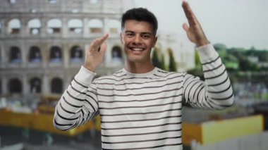 Roma ikonik stadyumunun önünde neşeli bir şekilde poz veren genç adam, açık bir gülümseme ve çizgili bir gömlek sergiliyor, canlı bir şehir atmosferini yakalıyor..