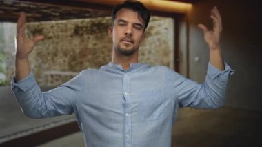 İçerideki İspanyol genç adam neşeli bir ifadeyle odada komik jestler yapıyor ve açık mavi bir gömlek giyiyor, doğal ışıklandırmada espri yapıyor..