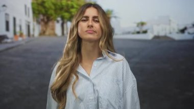 Genç sarışın kadın sağ eliyle avuç içi silkiyor ve köy caddesinde kaşlarını sarıyor; kararsızlık rahatsızlığı.