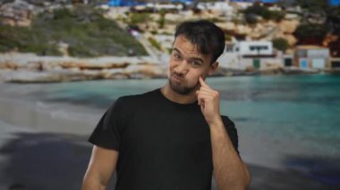 Sahil kenarında gülümseyen genç bir adam berrak mavi sularda ve arka planda kayalık kayalıklarla kumsalda güneşli bir günün tadını çıkarıyor, açık havada rahat bir yaşam tarzı sergiliyor..