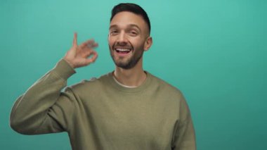Sıradan bir süveter içinde gülümseyen genç bir adam canlı mavi bir arka plana karşı olumlu ve kendine güvenen bir jest yapar..