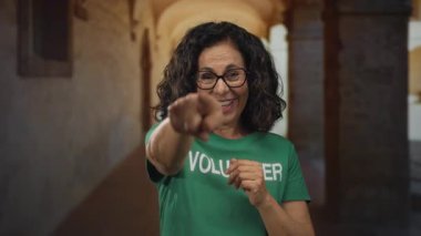 Orta yaşlı İspanyol kadın gönüllü tişörtlü, eski bir şehir caddesinde coşkuyla gülümseyen ve el kol hareketi yapan.