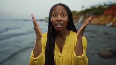 Sahilde dikilen, sarı tişörtlü, saçları örgülü kayalık sahil şeridine karşı duygularını ifade eden ve bulutlu gökyüzünün altında dalgalanan bir kadın..