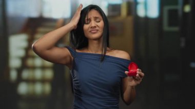 Otelde elinde nişan yüzüğü olan bir kadın mavi elbise giymiş, kapalı bir yerde gün ışığında genç ve düşünceli bir şekilde modern iç dekorda, İspanyol asıllı, zarif bir tavırla ortaya çıkıyor..