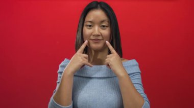 Canlı kırmızı bir duvarın önünde parmakları yanaklarında gülümseyen bir kadın, açık mavi süveter giyen, izole edilmiş arka plan, neşe ve pozitiflik gösteren genç bir kadın..