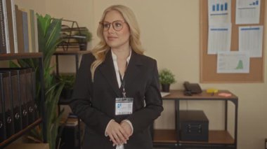 Ofis ortamında gülümseyen bir kadın. Profesyonel tavırları, sarı saçları ve gözlükleri var. Ofis ekipmanları ve evrakları arasında güvenle duruyor..