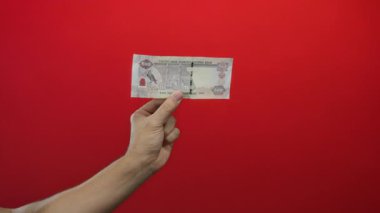 Kafkasyalı bir adam Birleşik Arap Emirlikleri 'ne bağlı Dirham Dirham banknotunu canlı kırmızı bir arka plana karşı tutuyor, izole ve odaklanmış bir şekilde para gösteriyor..