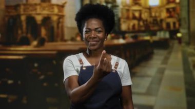 Önlüklü kadın kilise ortamında parmaklarıyla jest yapıyor, geleneği ve çağdaş stili ev ortamında güven ve cazibeyle harmanlıyor..