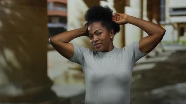 Güneşli bir günde afro saç stiliyle parkta gülümseyen, günlük kıyafetlerle dışarıda eğlenen, doğal güzelliğini ve mutluluğunu şehir ortamında sergileyen bir kadın..