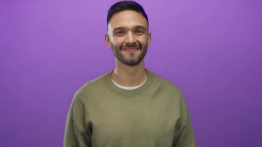 Genç İspanyol adam yeşil bir kazak giyerken canlı mor bir arka planın önünde gülümsüyor ve el sallıyor.