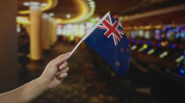 Elinde canlı kumarhane ortamında yeni Zelanda bayrağı tutan ve arka planda ışıklı kumar makineleri olan adam, kapalı alanlarda vatanseverliği vurguluyor..