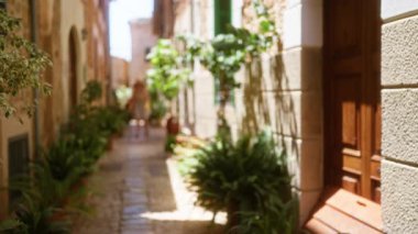 Bulanık bir kadın Mallorca 'da dar bir sokakta yürüyor, taş binalar ve parlak güneş ışığı altındaki yol boyunca canlı yeşil bitkiler rüya gibi, sıcak bir atmosfer yaratıyor..