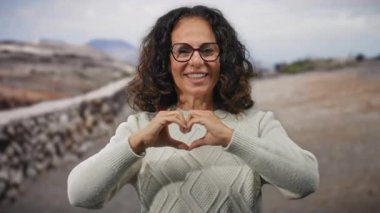 Bahçede güneşli bir günde, güzel kıvırcık saçları ve gözlükleriyle kalp jesti yapan bir kadın, açık hava parkında elleri ile mutluluk ve sıcaklık gösterisi yapıyor..